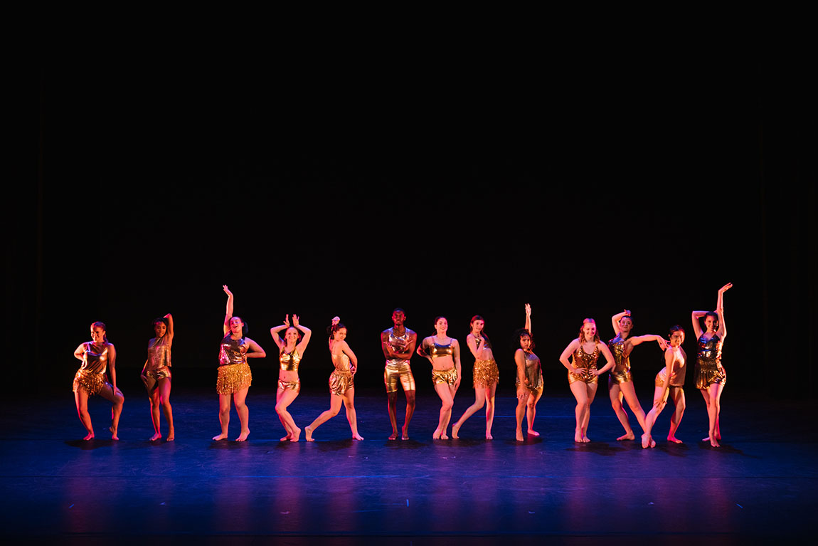 A row of college student dancers in shiny gold outfits strike a variety of poses in a line across the stage